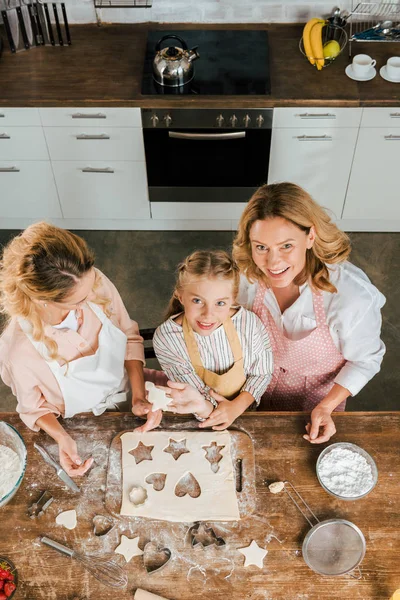 Vue du dessus de l'enfant heureux avec la mère et la grand-mère préparant des biscuits de Noël à la maison et regardant la caméra — Photo de stock