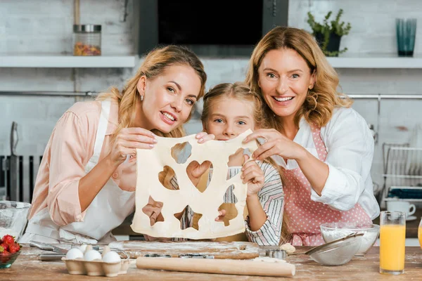 Happy child with mother and grandmother holding cutted dough with heart, star and christmas tree shaped holes at home — Stock Photo