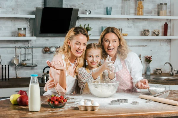 Feliz niño aplaudiendo las manos con harina mientras cocina con la madre y la abuela en casa - foto de stock