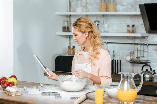 Beautiful young woman making dough and using tablet at kitchen — Stock Photo