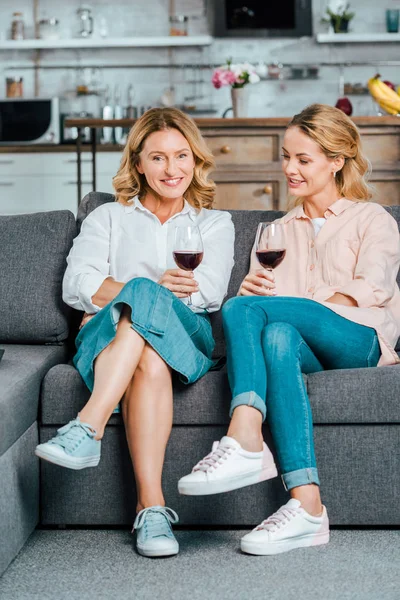 Зріла мати і доросла дочка в келихах червоного вина проводять час разом на дивані вдома — стокове фото