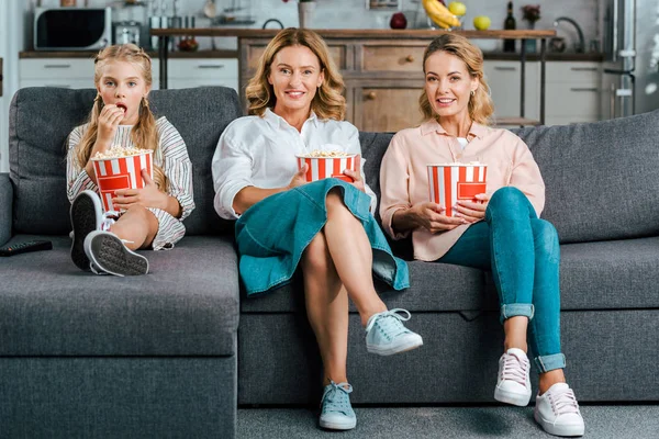 Счастливый ребенок с мамой и бабушкой смотреть кино на диване дома с ведрами попкорна — стоковое фото