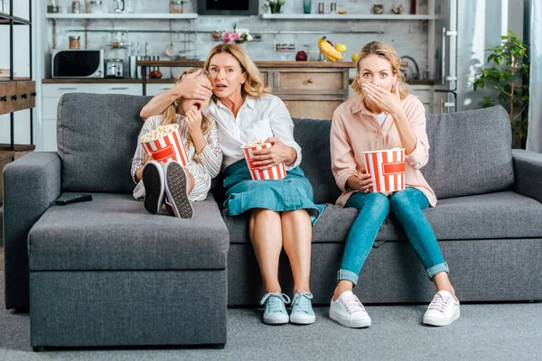 Испуганный ребенок с мамой и бабушкой, смотрящими кино дома на диване с ведрами попкорна — стоковое фото