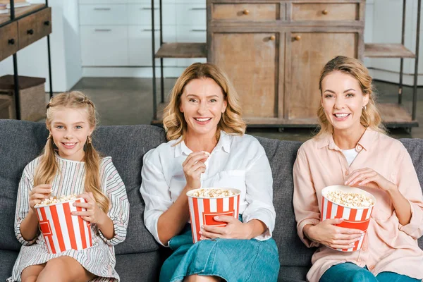 Happy child with mother and grandmother watching movie on couch at home with buckets of popcorn — Stock Photo