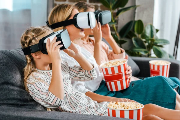 Visão lateral da criança com mãe e avó sentadas no sofá juntas e assistindo filme com fones de ouvido de realidade virtual em casa — Fotografia de Stock