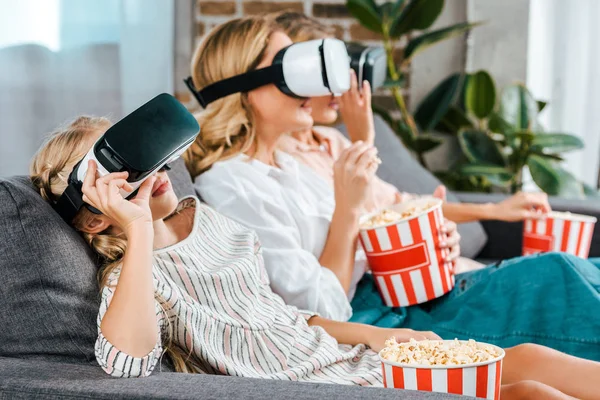 Pequena criança com mãe e avó sentados no sofá juntos e assistindo filme com fones de ouvido realidade virtual em casa — Fotografia de Stock