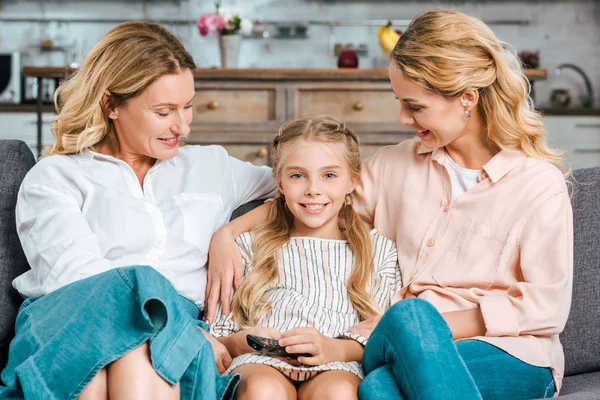 Child with mother and grandmother sitting on couch together with remote control at home — Stock Photo