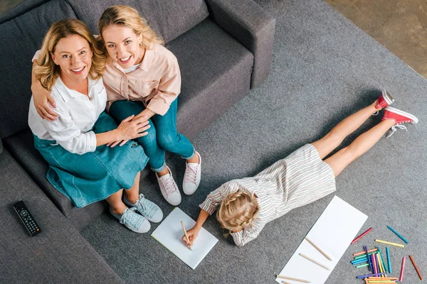 Vue grand angle de mère et grand-mère heureux assis sur le canapé tandis que l'enfant dessin sur le sol à la maison — Photo de stock