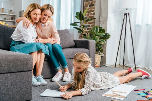 Glückliche Mutter und Großmutter auf dem Sofa sitzend, während das Kind zu Hause auf dem Fußboden zeichnet — Stockfoto