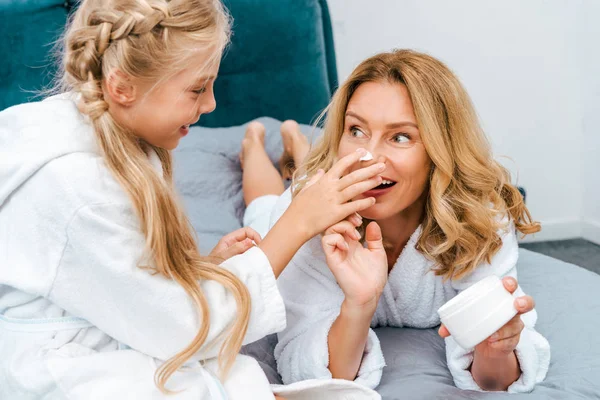 Mãe feliz e filha em roupões relaxando na cama e aplicando creme facial — Fotografia de Stock