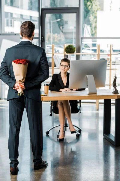 Vista trasera del hombre de negocios escondiendo ramo de rosas detrás de la espalda para sorprender a la mujer de negocios en la oficina - foto de stock