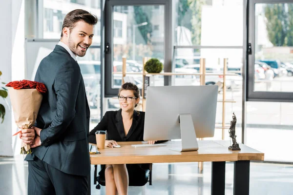 Sonriente hombre de negocios escondiendo ramo de rosas detrás de la espalda para sorprender a la mujer de negocios en el cargo - foto de stock