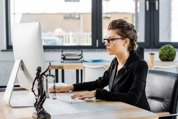 Attraente donna d'affari in occhiali che lavora con il computer a tavola in ufficio, themis statua sul tavolo — Foto stock