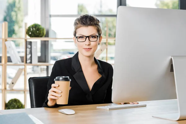 Attractive businesswoman in glasses working with computer at table in office and holding disposable coffee cup — Stock Photo