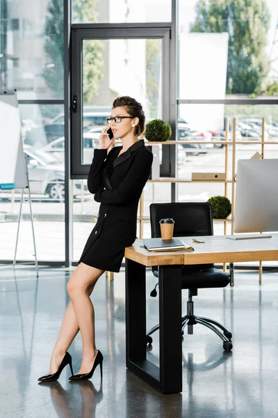 Seitenansicht einer attraktiven Geschäftsfrau, die sich im Büro auf den Tisch lehnt und mit dem Smartphone spricht — Stockfoto
