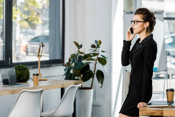 Вид збоку красивої бізнес-леді в формальному одязі, спираючись на стіл і розмовляючи за допомогою смартфона в офісі — стокове фото