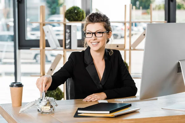 Sonriente atractiva mujer de negocios poner dinero en frasco de vidrio con nota Vacaciones en la oficina, concepto de viaje - foto de stock