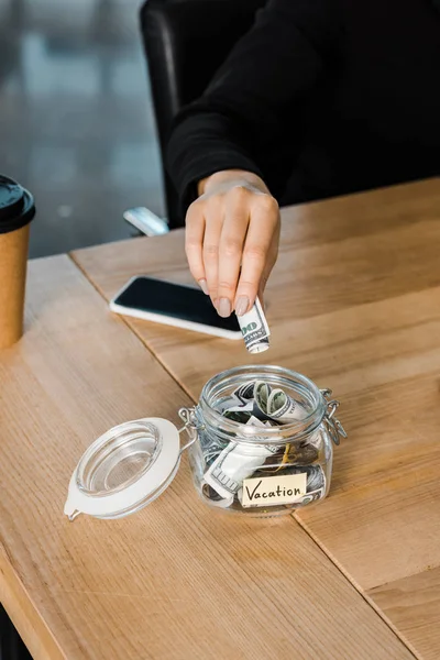 Cropped image of businesswoman putting dollar banknotes in glass jar with note Vacation in office — Stock Photo