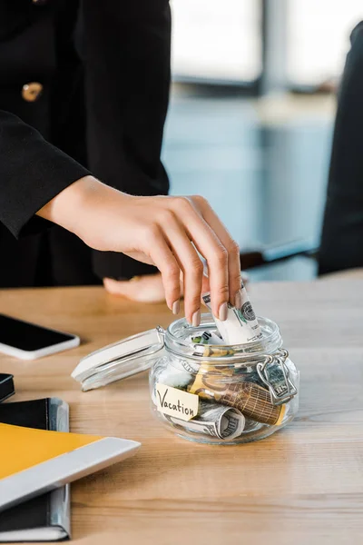 Imagem cortada de empresária colocando dinheiro em frasco de vidro com nota Férias no escritório, conceito de viagem — Fotografia de Stock