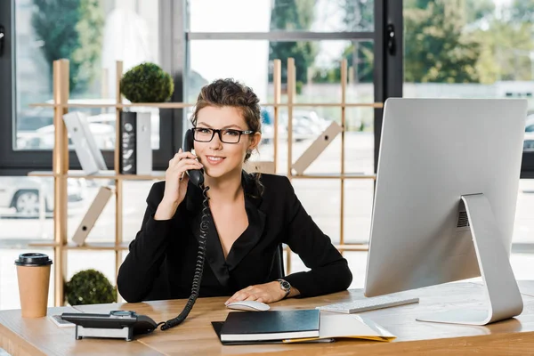 Улыбающаяся привлекательная деловая женщина разговаривает по стационарному телефону в офисе и смотрит в камеру — стоковое фото