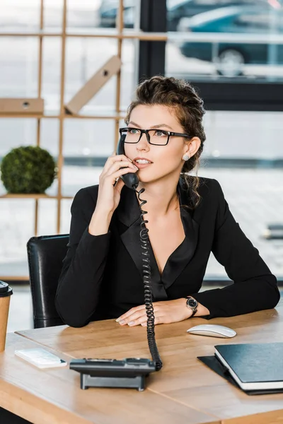 Attractive businesswoman talking by stationary telephone in office and looking away — Stock Photo