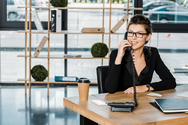 Smiling beautiful businesswoman talking by stationary telephone in office and looking at camera — Stock Photo