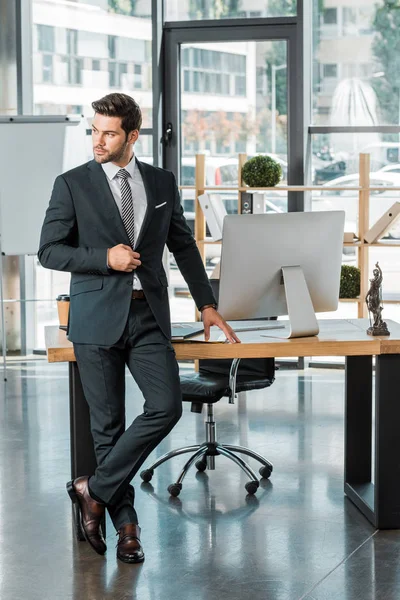 Hombre de negocios guapo de pie cerca de la mesa en la oficina, tocando la chaqueta y mirando hacia otro lado - foto de stock