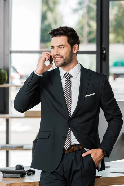 Усміхнений красивий бізнесмен, спираючись на стіл і розмовляючи по смартфону в офісі — стокове фото