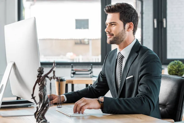 Schöner Anwalt arbeitet mit Computer am Tisch im Büro, themis Statue auf Tischplatte — Stockfoto