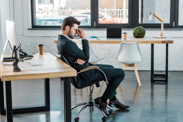 Vista lateral del hombre de negocios cansado sentado en la silla y tocar el puente de la nariz en la oficina - foto de stock