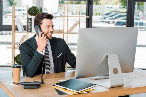 Sonriente hombre de negocios guapo hablando por teléfono fijo en la oficina y mirando el ordenador - foto de stock