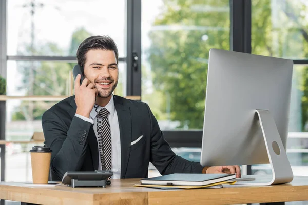 Smiling handsome businessman talking by stationary telephone in office and looking at camera — Stock Photo