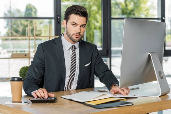 Hombre de negocios guapo usando calculadora en la oficina, carpeta abierta en la mesa - foto de stock