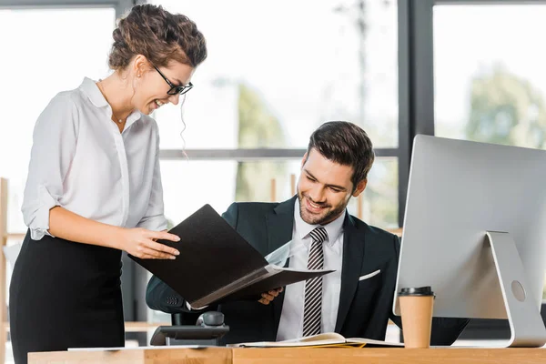 Sonriente secretaria mostrando carpeta con documentos a guapo hombre de negocios en la oficina - foto de stock