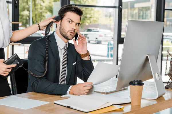 Sorprendido hombre de negocios hablando por teléfono inteligente y secretario sosteniendo el teléfono fijo cerca de su oído - foto de stock