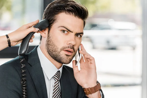 Shocked businessman talking by smartphone and secretary giving him handset of stationary telephone — Stock Photo