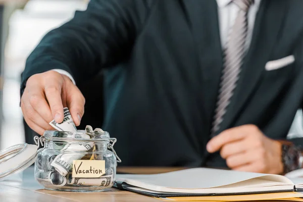 Cropped shot of businessman putting money into glass jar with vacation inscription at workplace — Stock Photo