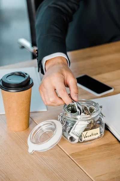 Обрізаний знімок бізнесмена покласти гроші в скляну банку з відпусткою напис на робочому місці — стокове фото