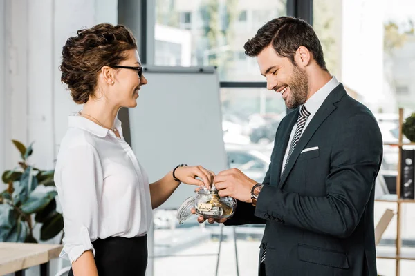 Side view of smiling business colleagues putting money into glass jar with vacation lettering in office — Stock Photo