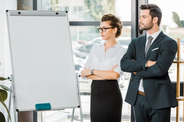 Portrait of young business colleagues with arms crossed standing at empty white board in office — Stock Photo