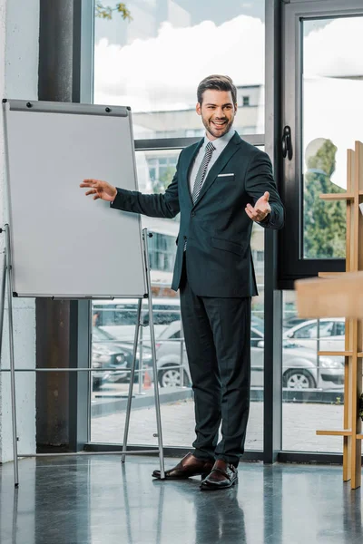 Молодий усміхнений бізнесмен у костюмі, стоячи на порожній білій дошці в офісі — стокове фото