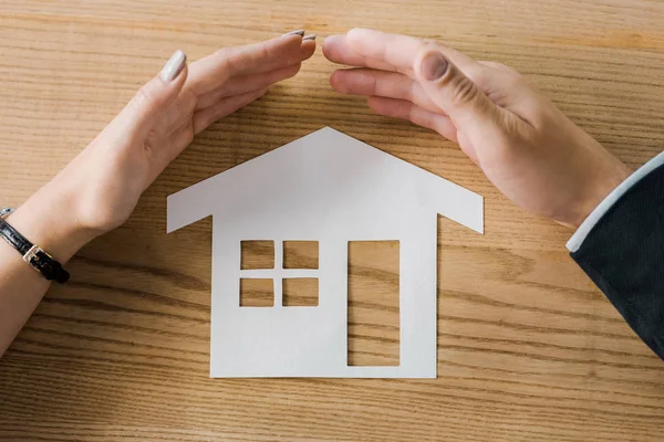 Partial view of business people covering house paper model on wooden tabletop with hands, insurance concept — Stock Photo