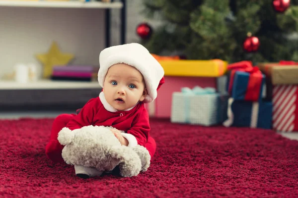 Крупним планом портрет красивої маленької дитини в костюмі Санти лежить на червоному килимі з плюшевим ведмедем перед різдвяною ялинкою і подарунками і дивиться на камеру — стокове фото