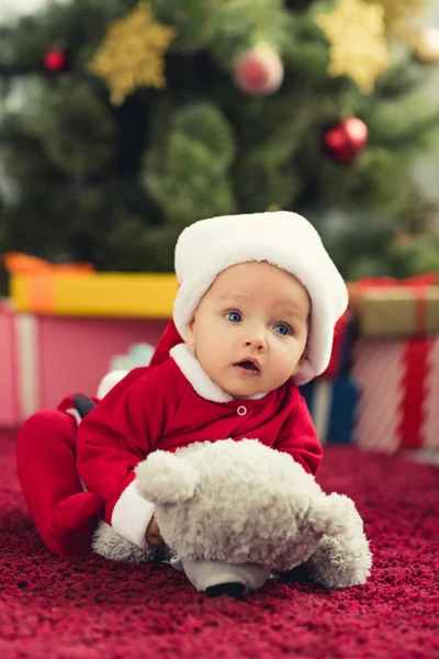 Close-up portrait of cute little baby in santa suit lying on red carpet with teddy bear in front of christmas tree and gifts — Stock Photo