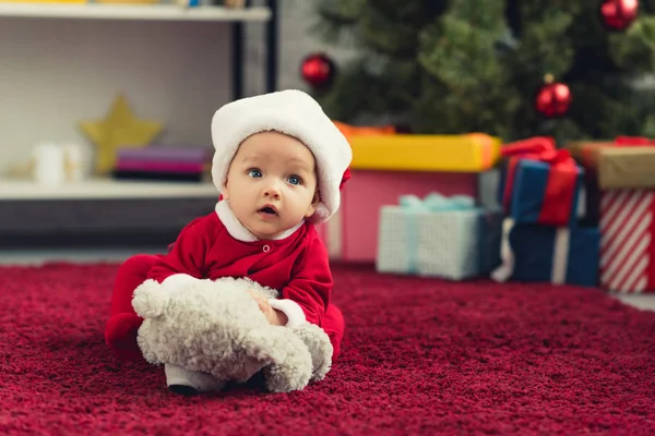 Retrato de primer plano de adorable bebé en traje de santa acostado en la alfombra roja con oso de peluche delante del árbol de Navidad y regalos - foto de stock