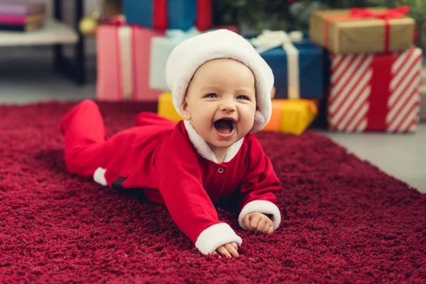 Ridendo piccolo bambino in costume da Babbo Natale sdraiato sul tappeto rosso davanti all'albero di Natale e regali — Foto stock