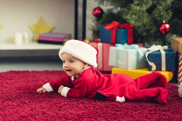 Adorable bebé en traje de santa tumbado en el suelo delante del árbol de Navidad y regalos - foto de stock