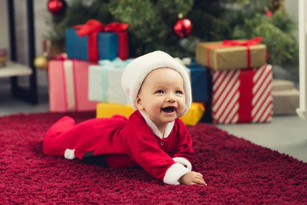 Laughing little baby in santa hat lying on floor in front of christmas tree and gifts — Stock Photo