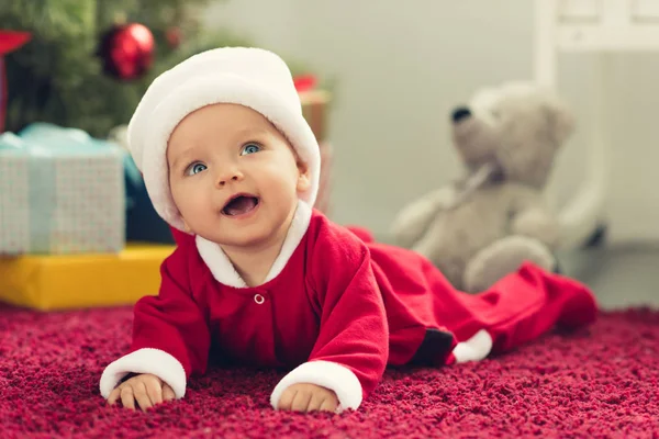 Ritratto ravvicinato di adorabile bambino in cappello di Babbo Natale con albero di Natale sfocato e regali sullo sfondo e guardando in alto — Foto stock