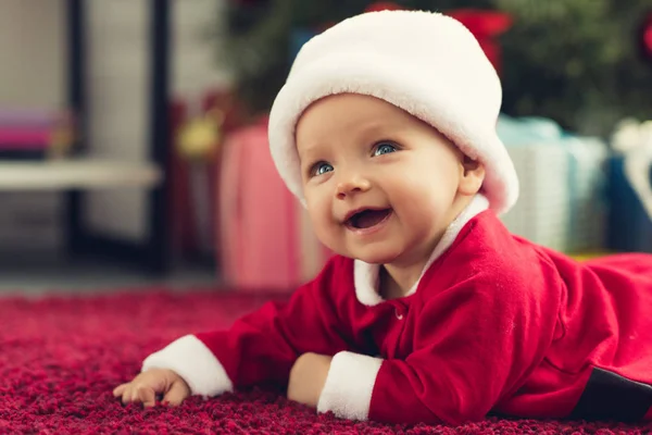 Happy little baby in santa suit lying on red carpet with christmas gifts blurred on background — Stock Photo